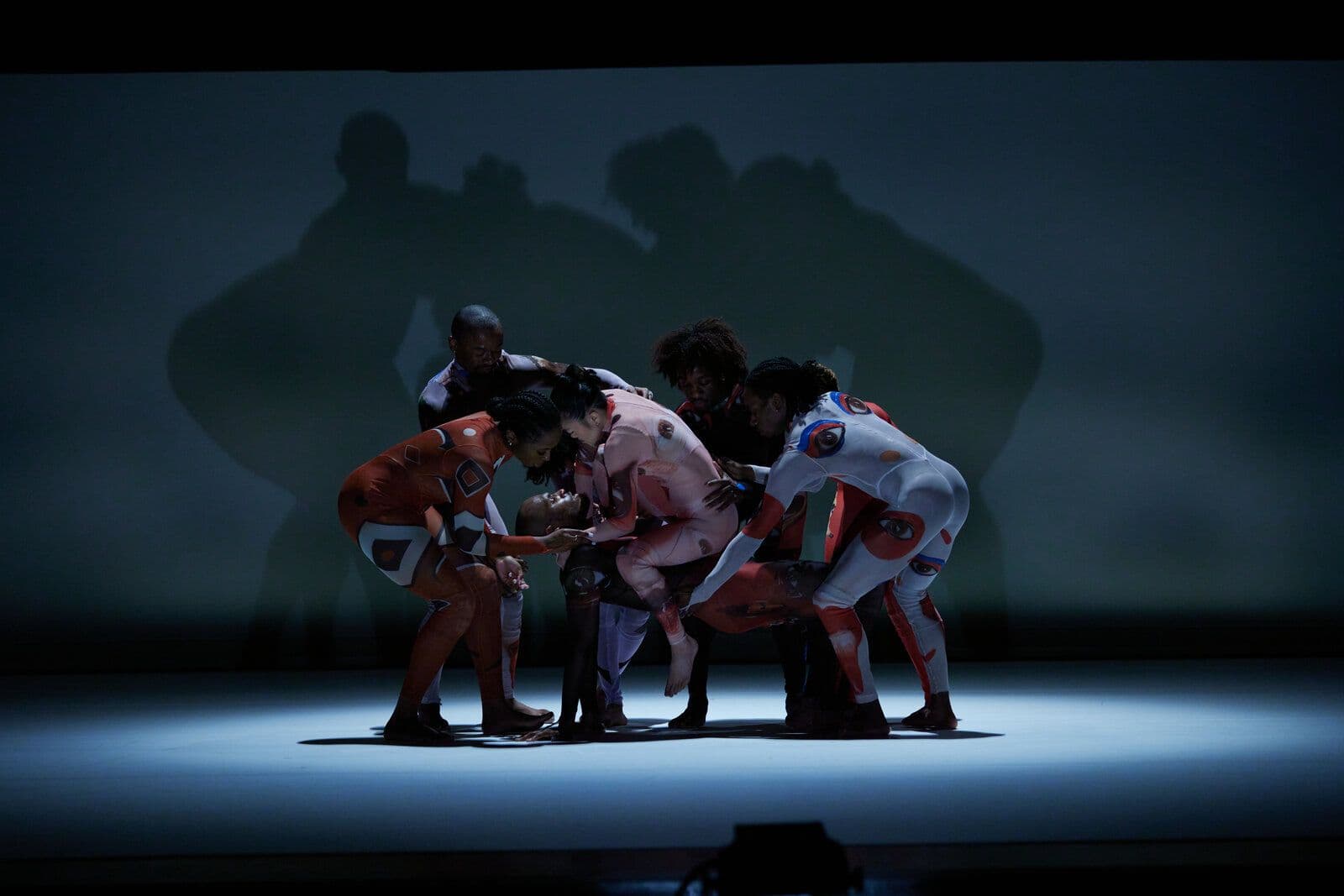 A group of dancers in patterned bodysuits huddle closely together, several bending in to gently hold and support a dancer lifted horizontally at the center, their combined shadows cast large on the backdrop under a dim spotlight.