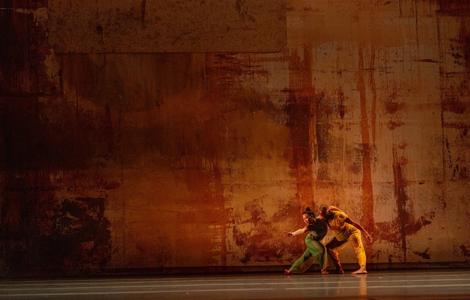 Dancers perform '2x4' onstage all lit in warm amber tones against a weathered rust-toned backdrop.