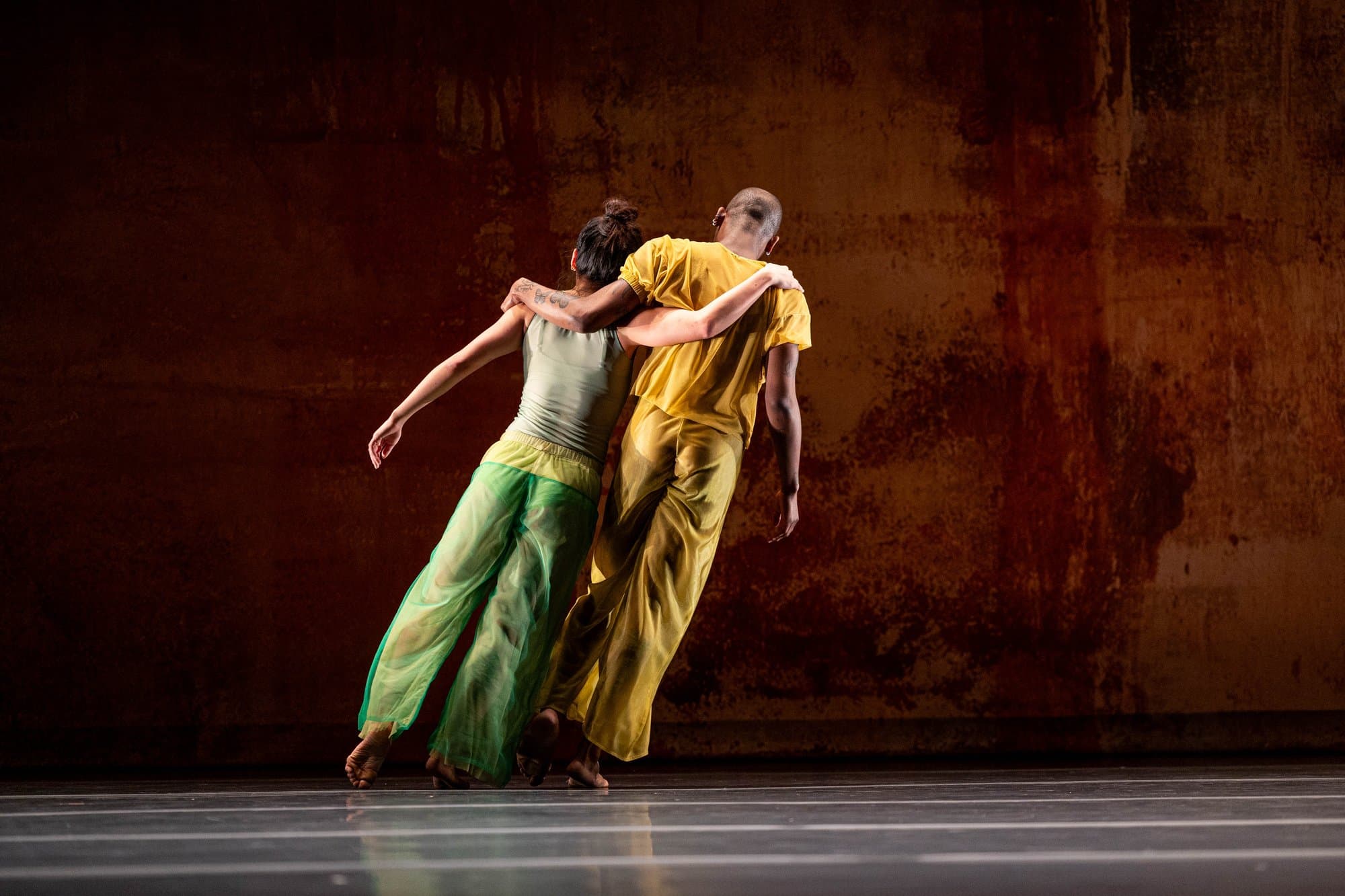 Two dancers, one in a green outfit and one in yellow, lean off-balance together in a low, weighted duet against a rust-colored textured backdrop, from '2x4' by Kyle Abraham