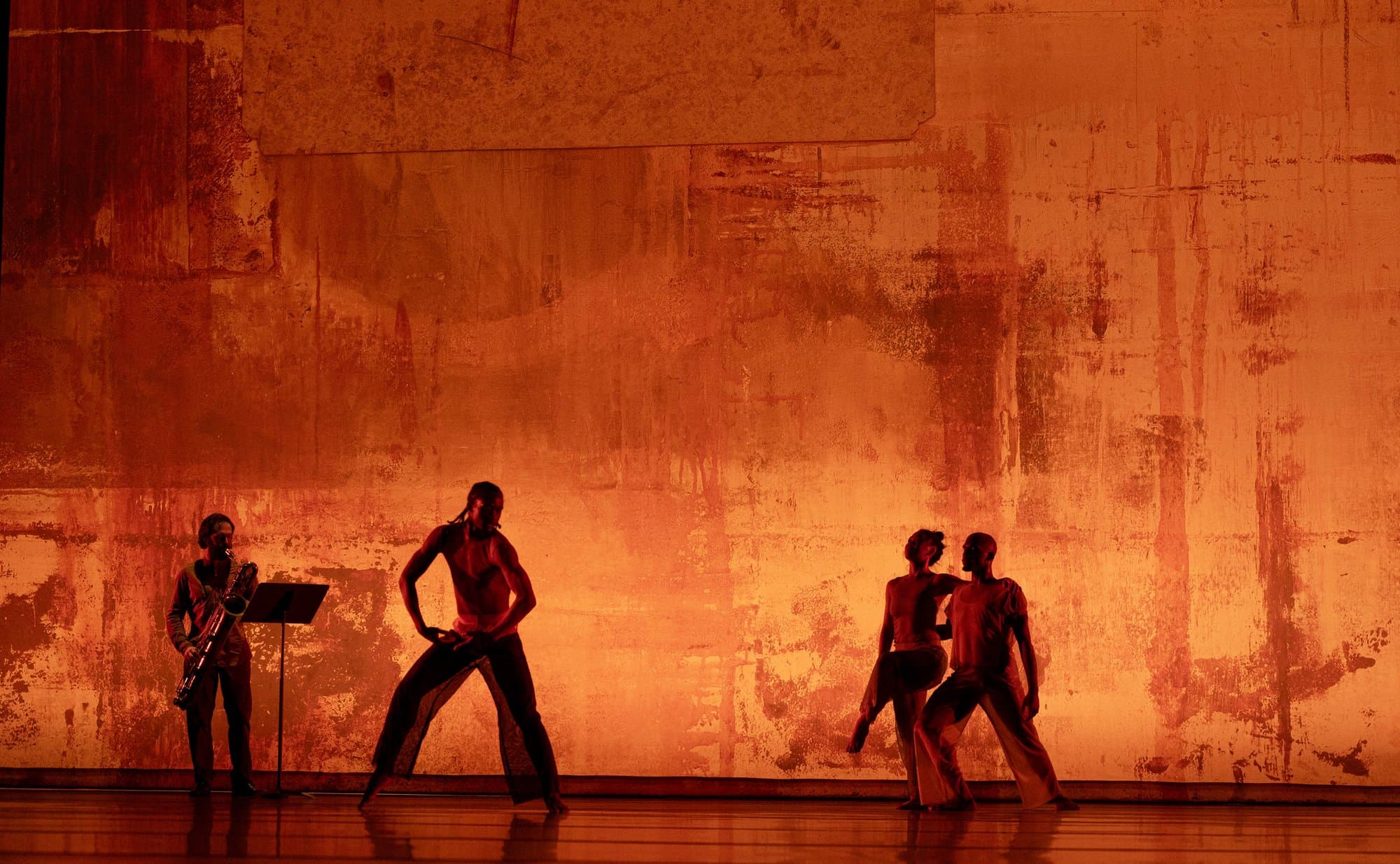 Dancers perform '2x4' onstage: a shirtless dancer in a wide lunge in the foreground, a live saxophonist at a music stand on the left, and two dancers in a close duet on the right, all lit in warm amber tones against a weathered rust-toned backdrop.
