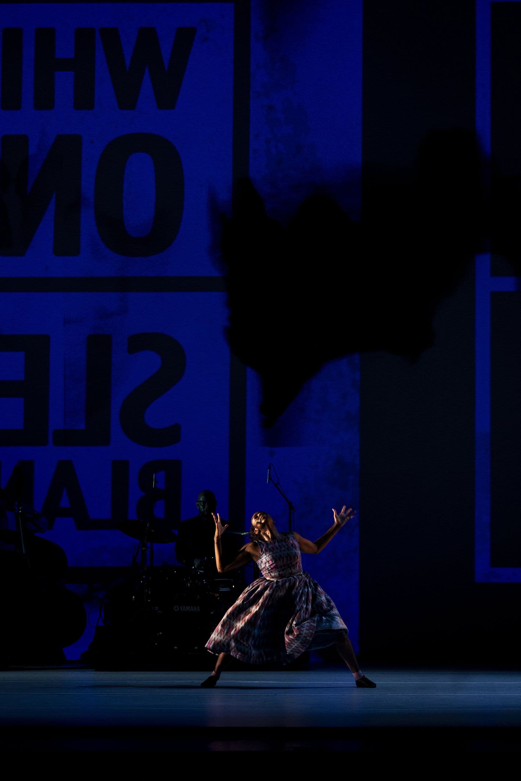 A dancer in a floral dress lunges dramatically under blue stage lighting, arms raised, with a musician and large mirrored text visible in the background, from 'The Gettin'.