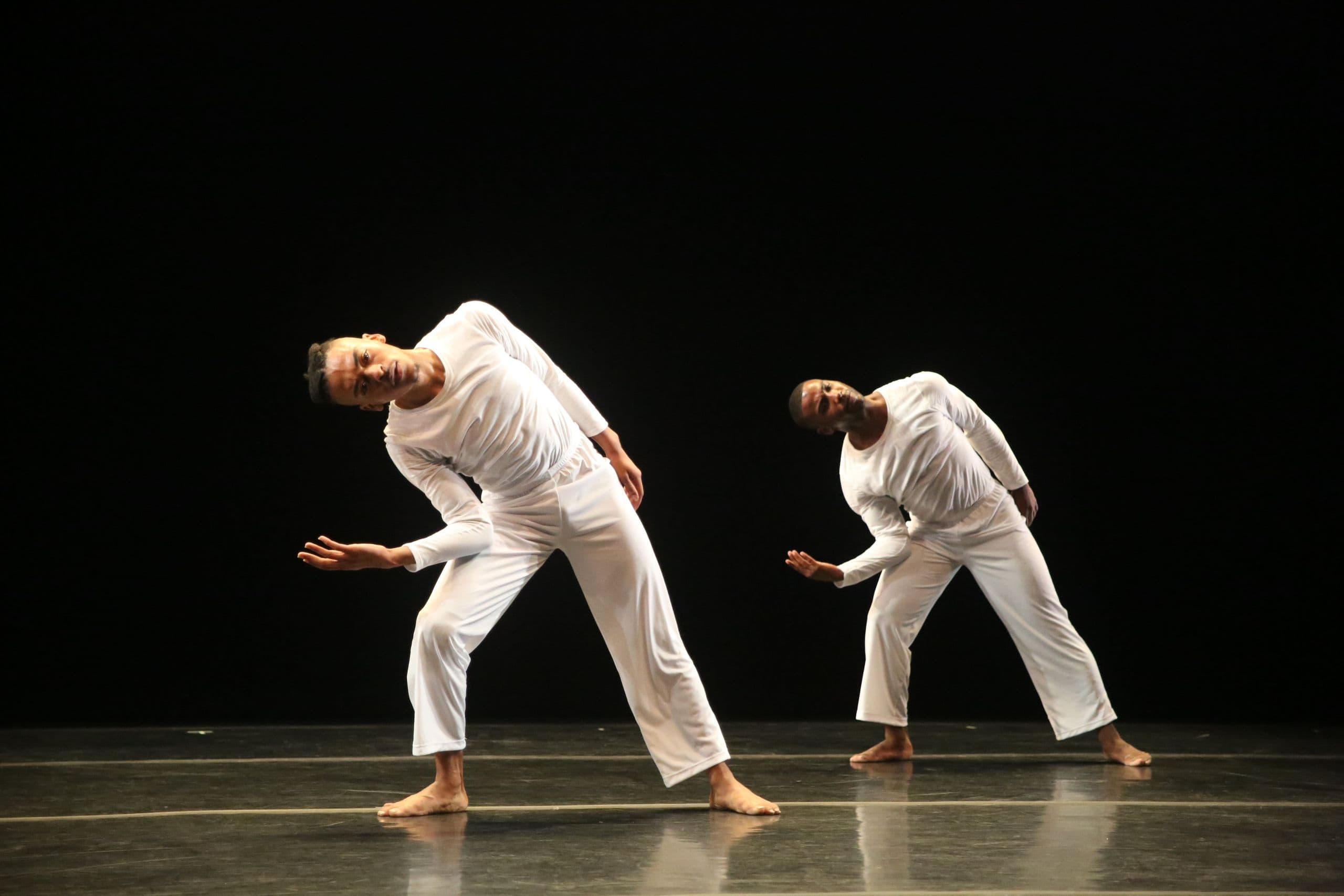 Two dancers in white lean sideways with arms extended on a dark stage, from 'Solo Olos.