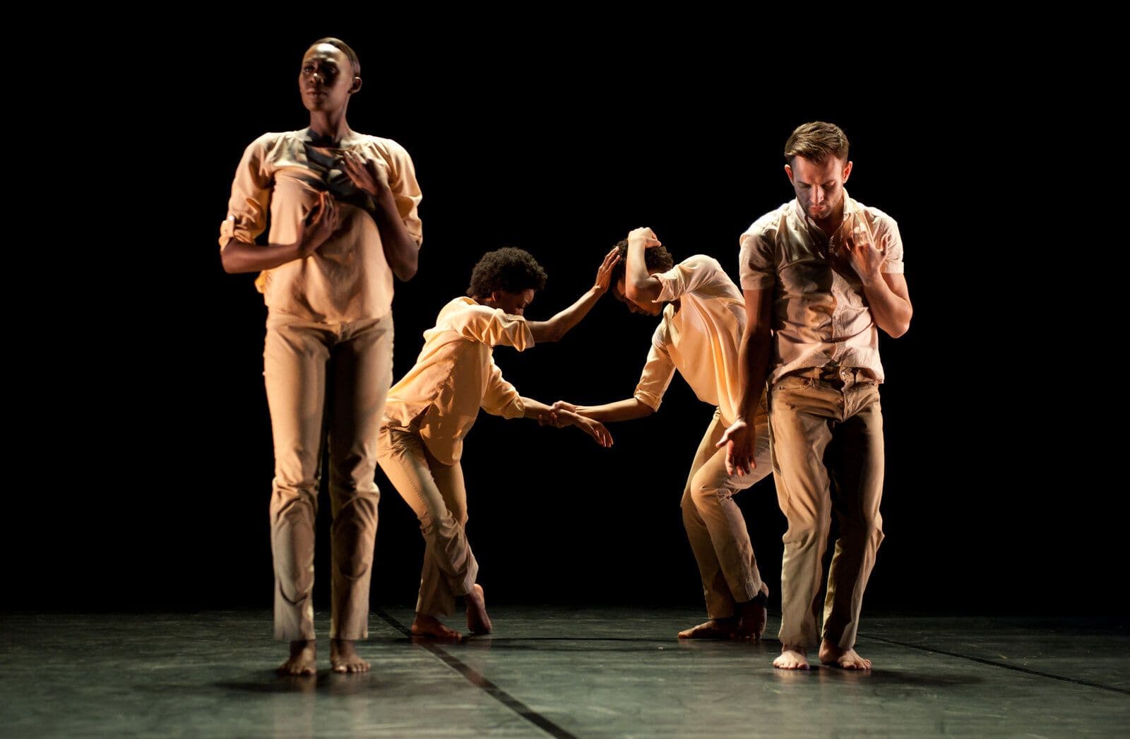 Four dancers in soft peach-toned clothing move through a chain of contact, one reaching to cradle another's head while two more stand apart, lit warmly against a black backdrop.