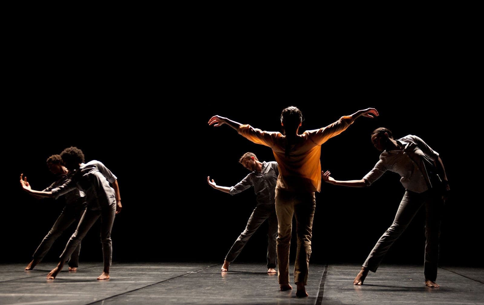 Six dancers move together on a dark stage, several bent forward with arms extended low while a central figure in an orange top stands with arms spread wide overhead.