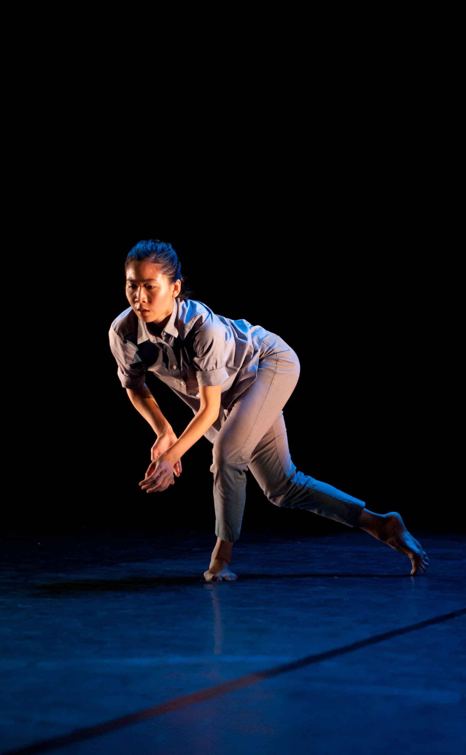 Dancer Connie Shiau leans low into a deep lunge with clasped hands, one leg extended far behind her, lit warmly against a dark blue-toned stage.