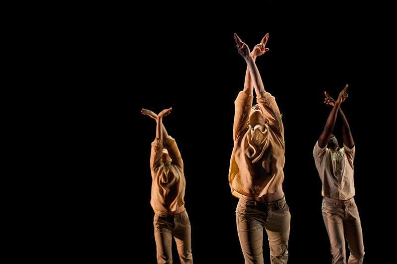 Three dancers reach both arms overhead with crossed wrists, caught mid-motion in matching golden-toned tops against a black backdrop.