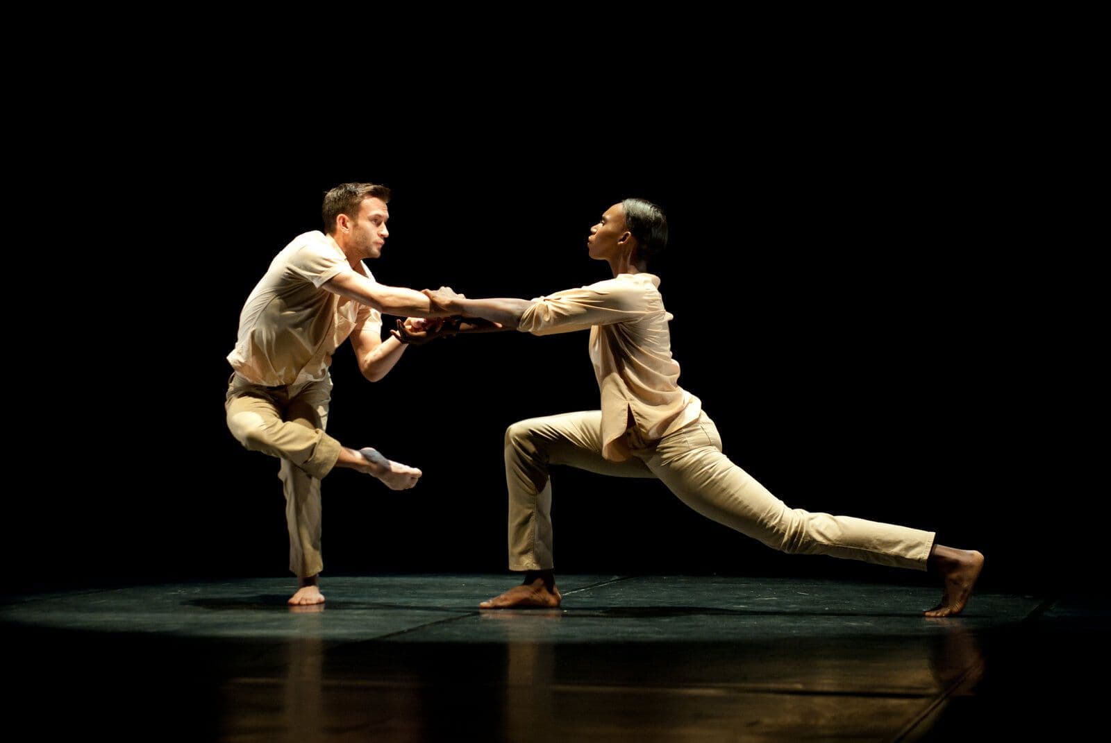 Dancers Matthew Baker and Penda N'diaye face each other in matching cream outfits, each in a low lunge with hands clasped between them, lit warmly against a dark stage.
