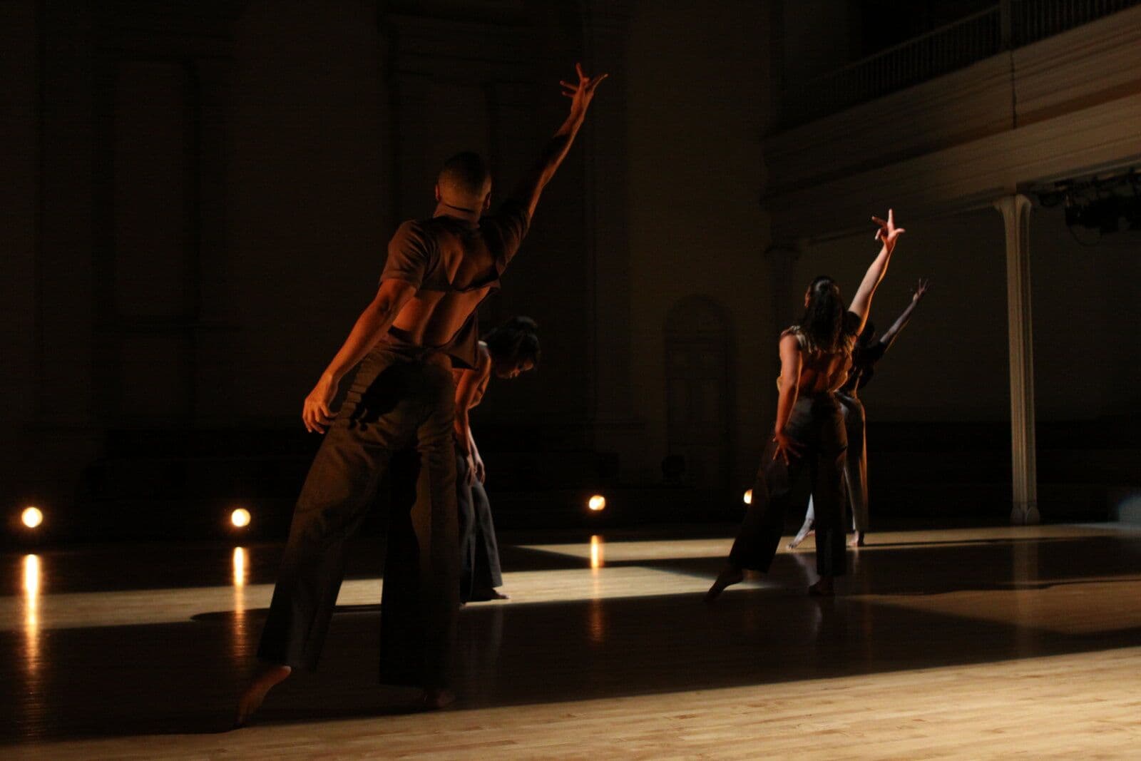 Four dancers in earth-toned clothing perform in a dimly lit grand hall, several reaching one arm skyward, illuminated by warm spotlights and small floor lights against tall archways in the background.