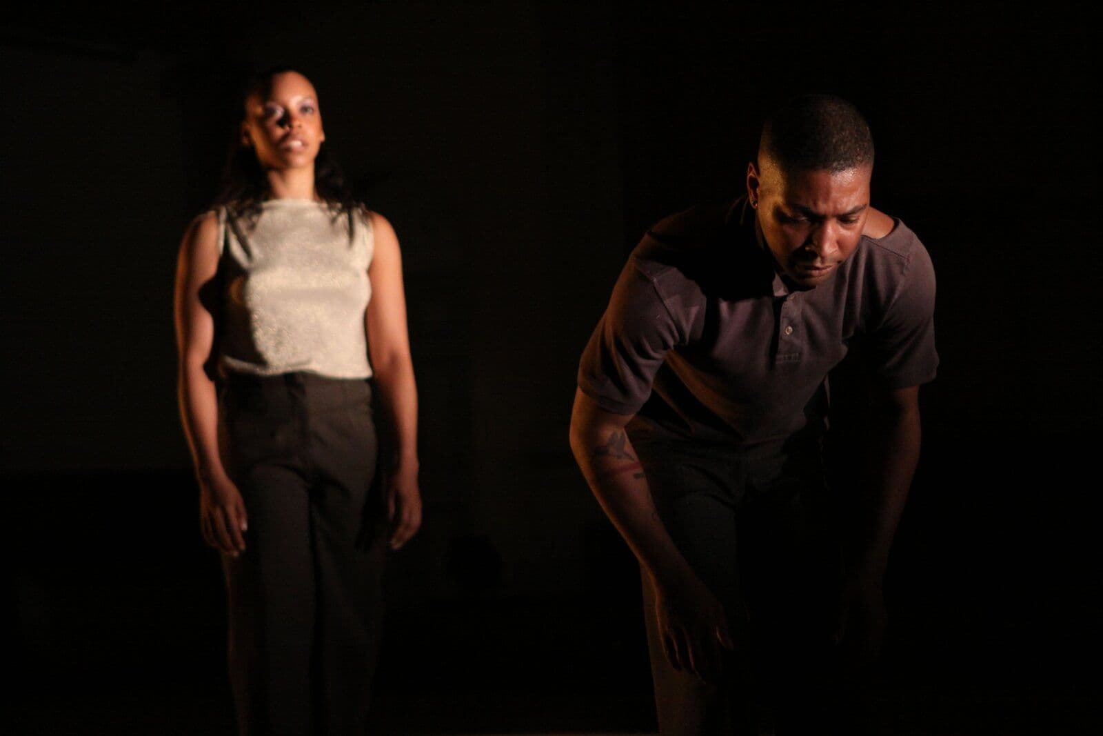Dancers Amber Lee Parker and Kyle Abraham perform apart in a dark space, she standing still and gazing down while he bends forward in a low, contained pose, both lit by warm spotlight against near-total darkness.