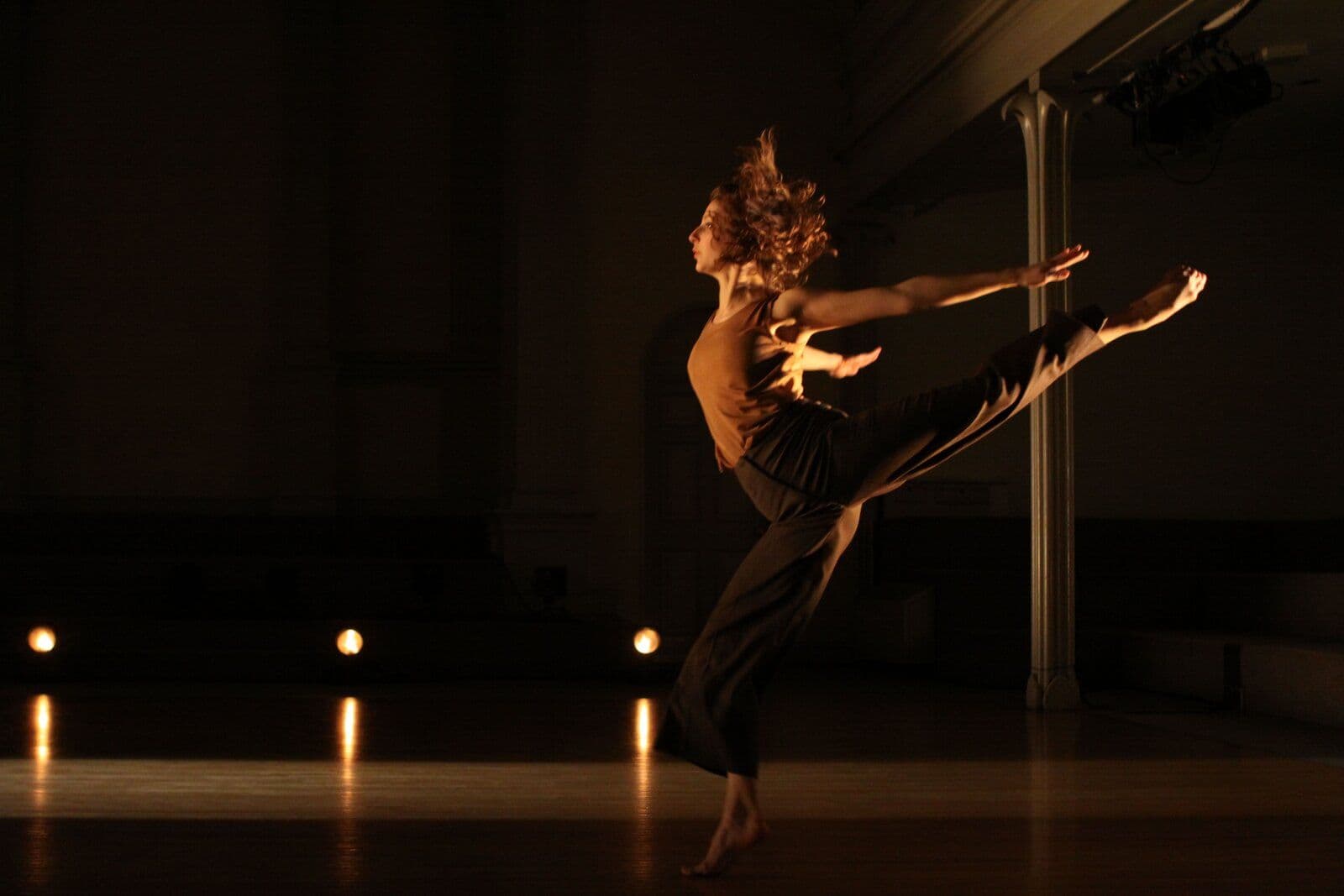 Dancer Rachelle Rafailedes leaps in profile with one leg extended high behind her and arms stretched forward, hair flying, lit warmly against a dark, columned interior.