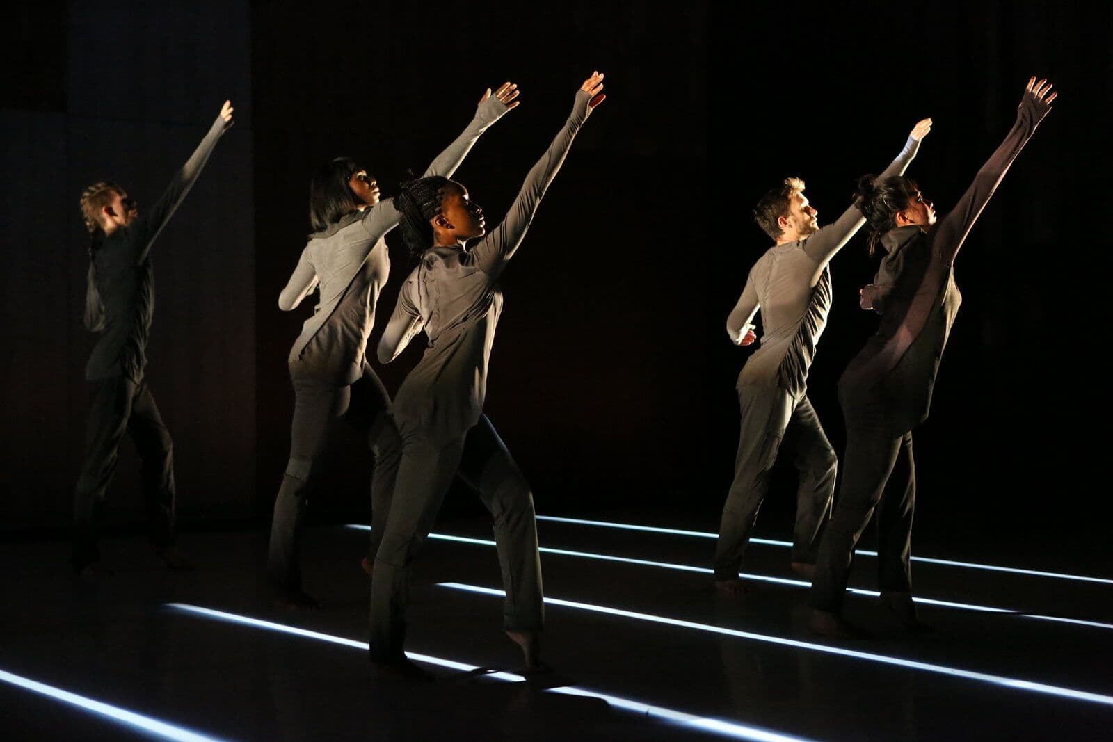 Six dancers in matching gray outfits reach one arm skyward in unison, lit by two glowing white light lines on the floor against a dark stage.
