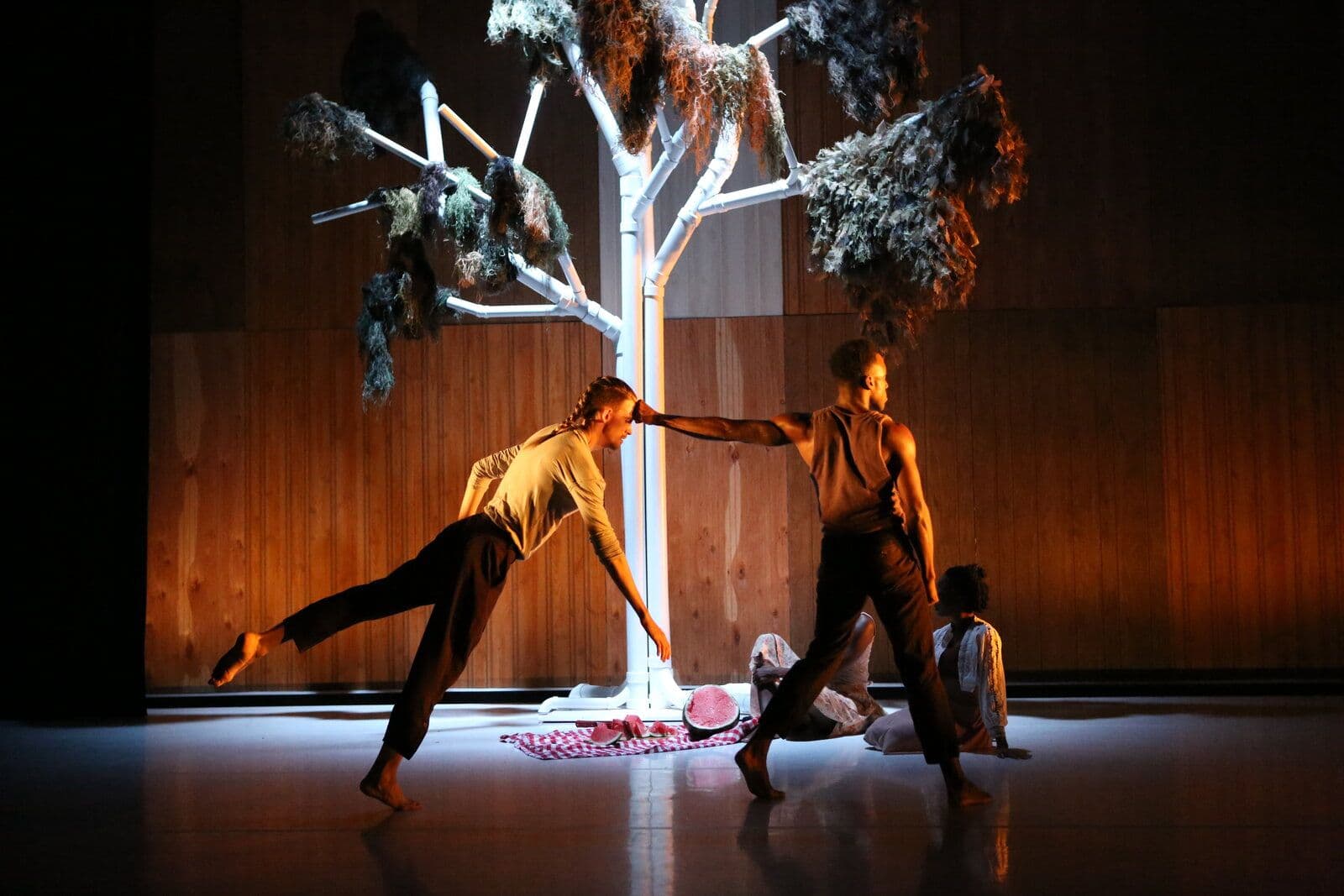 Two dancers reach toward each other beneath a large sculptural tree draped in moss, with a sliced watermelon on a checkered picnic blanket at its base and another performer seated nearby.