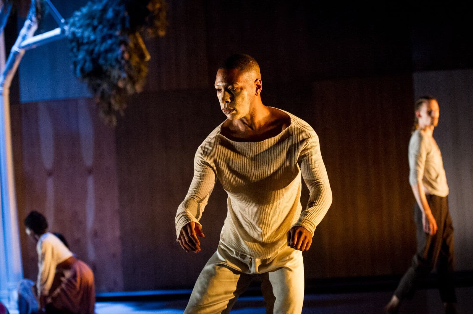 A dancer in a cream sweater and pants moves forward with fists loosely clenched, lit warmly beneath a sculptural tree hung with moss-like fabric, with other performers visible in the softly lit background.