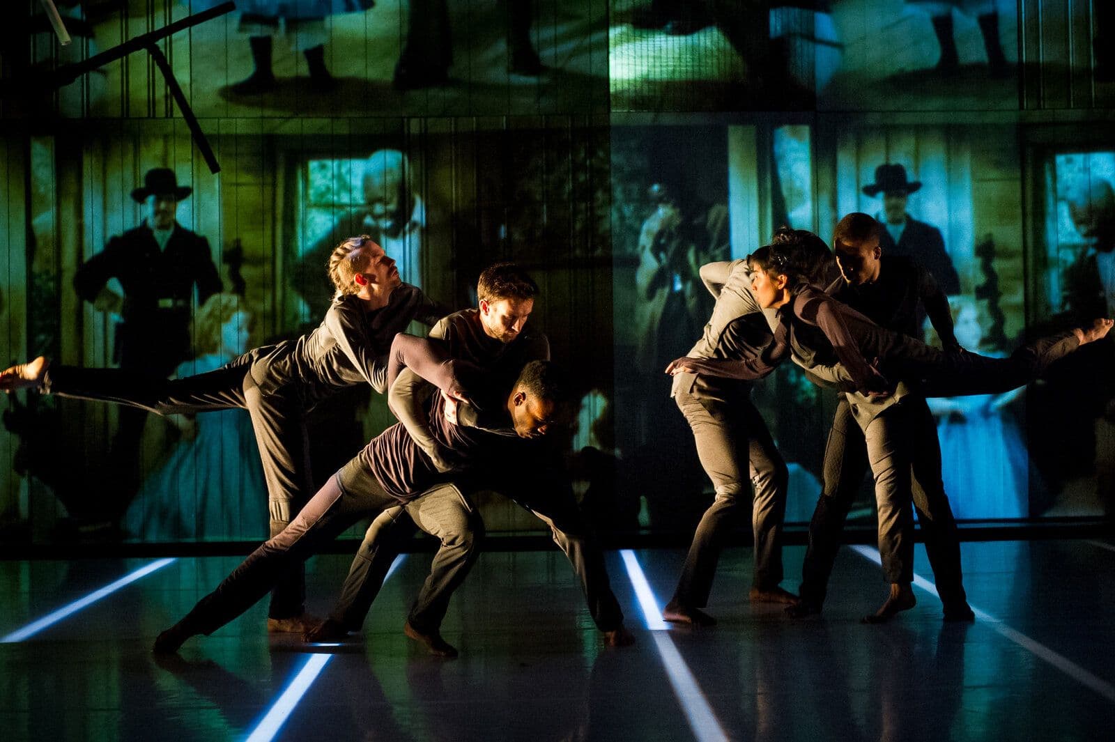 Six dancers in muted earth-toned clothing move in mirrored pairs, several extending a leg into a wide arabesque, in front of a large projected backdrop of vintage-style photographs.