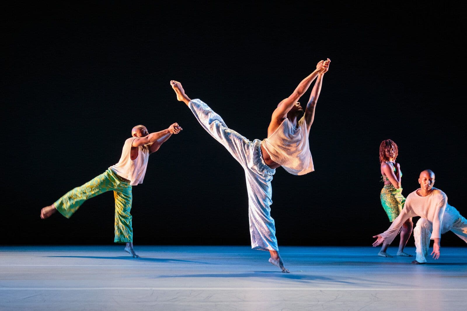 Two dancers in the foreground extend into a high side kick with clasped raised hands, wearing metallic and green patterned pants, while two more dancers move together in a low lunge in the background, on a dark stage.