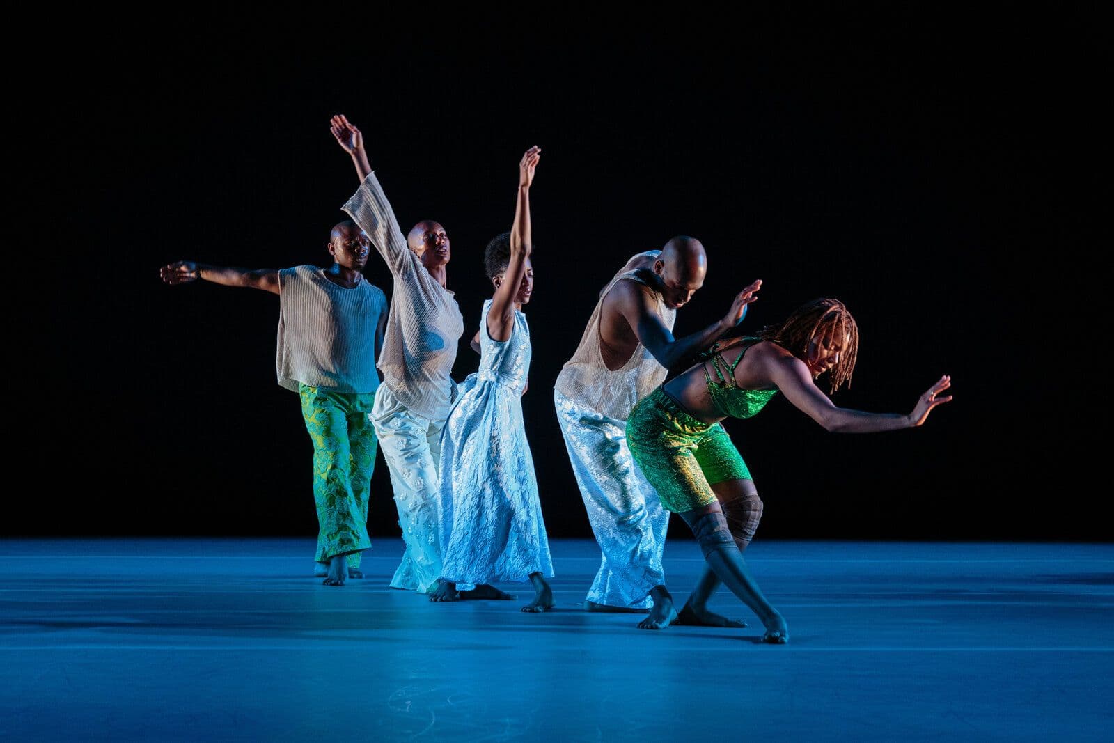 Five dancers in flowing white and shimmering green costumes move together across the stage, several with arms raised overhead and one leaning low with an arm extended, lit in cool blue tones against a black background.