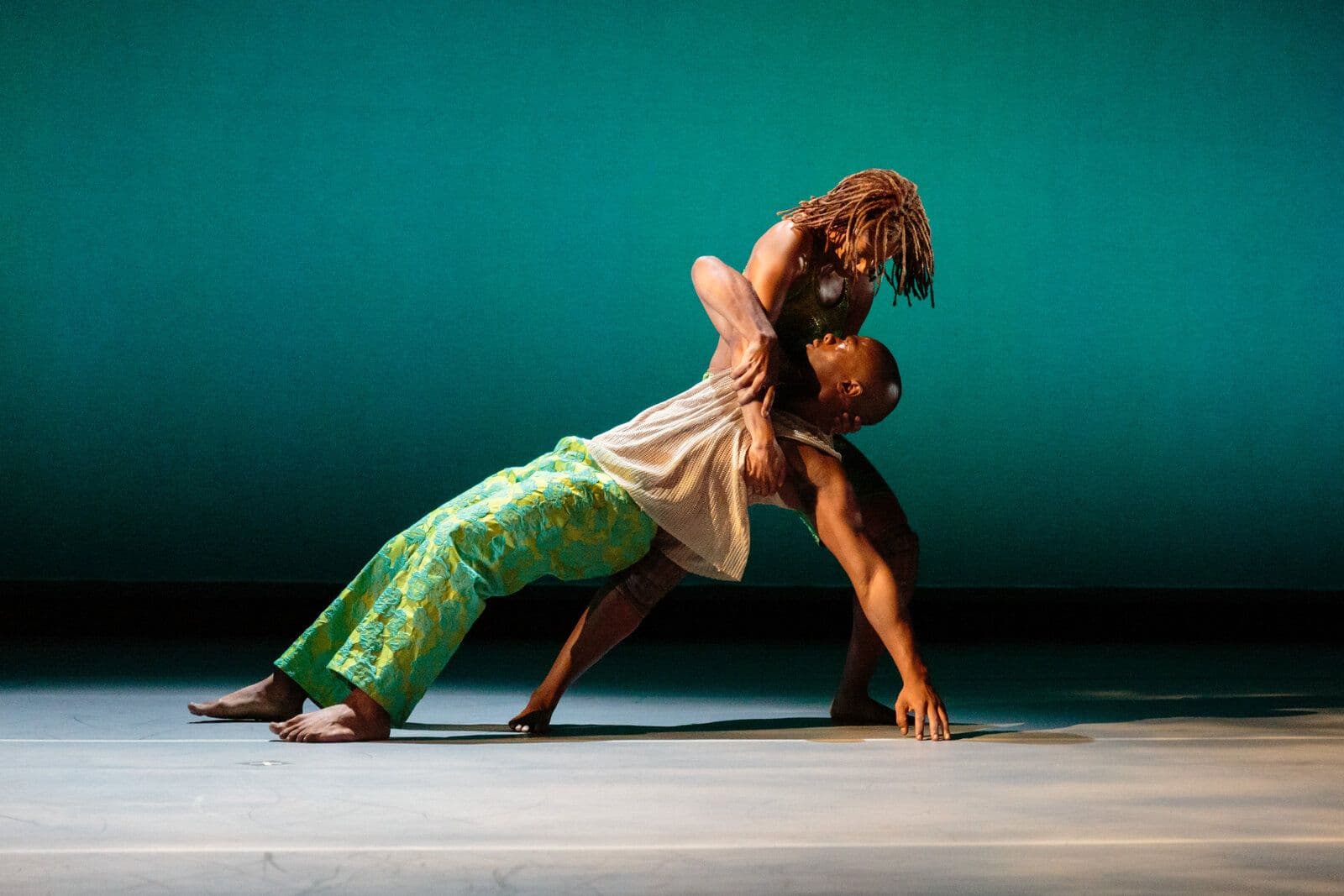 Two dancers embrace in a deep backbend, one leaning back with dreadlocks cascading down as the other supports her from below, set against a rich teal backdrop.
