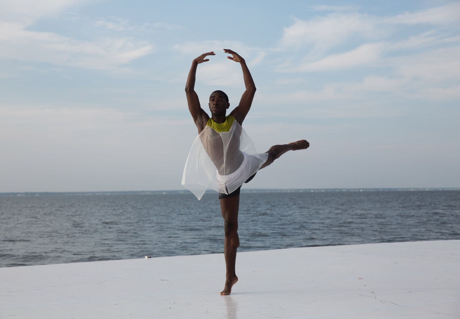 Dancer Chalvar Monteiro poses in an arabesque with arms raised overhead, wearing a sheer white and chartreuse costume, on a white outdoor platform against a coastal sky and open water.