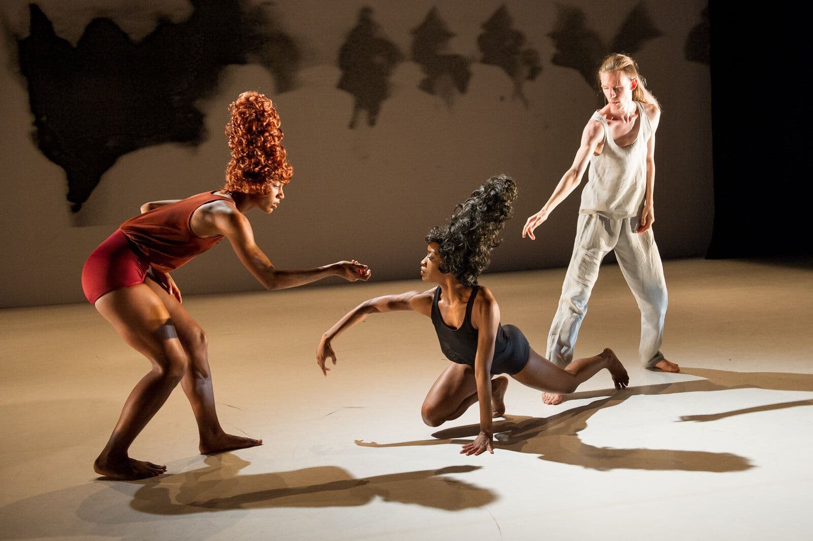Catherine Kirk, in a red top and tall red wig, reaches toward Tamisha A. Guy, who kneels low reaching back, while Jordan Morley in loose white clothing approaches from behind, all three lit warmly on a bare stage with shadowy silhouettes on the backdrop.