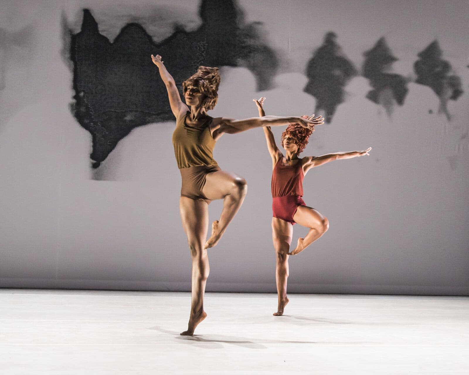 Two dancers in olive and maroon tops stand on one leg with the other knee bent and both arms raised in mirrored diagonal lines, performing in unison on a pale stage.