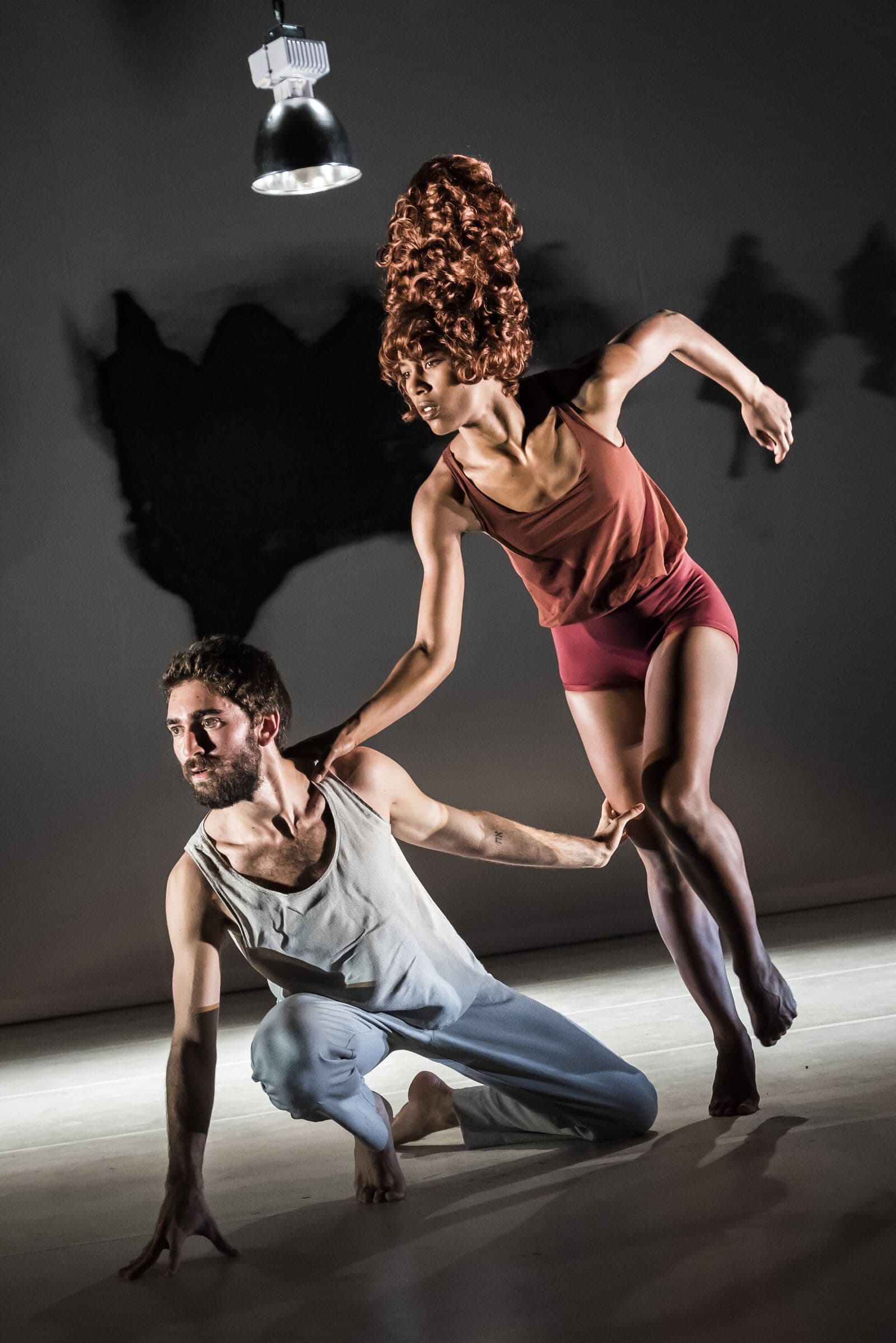 Sam Pratt kneels low on the floor while Catherine Kirk, wearing a tall curly red wig and rust-colored top, leaps beside him with one hand resting on his shoulder, lit dramatically under a single overhead light against a dark backdrop.