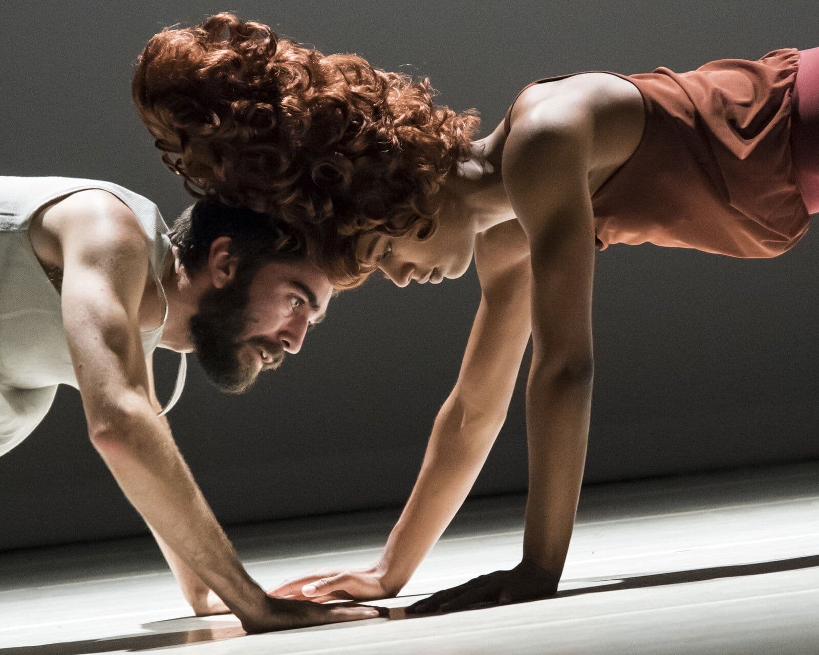 Sam Pratt and Catherine Kirk, her tall curly red wig framing his face, are captured in profile in matching low lunge poses with hands pressed to the floor, foreheads nearly touching, under a warm spotlight.