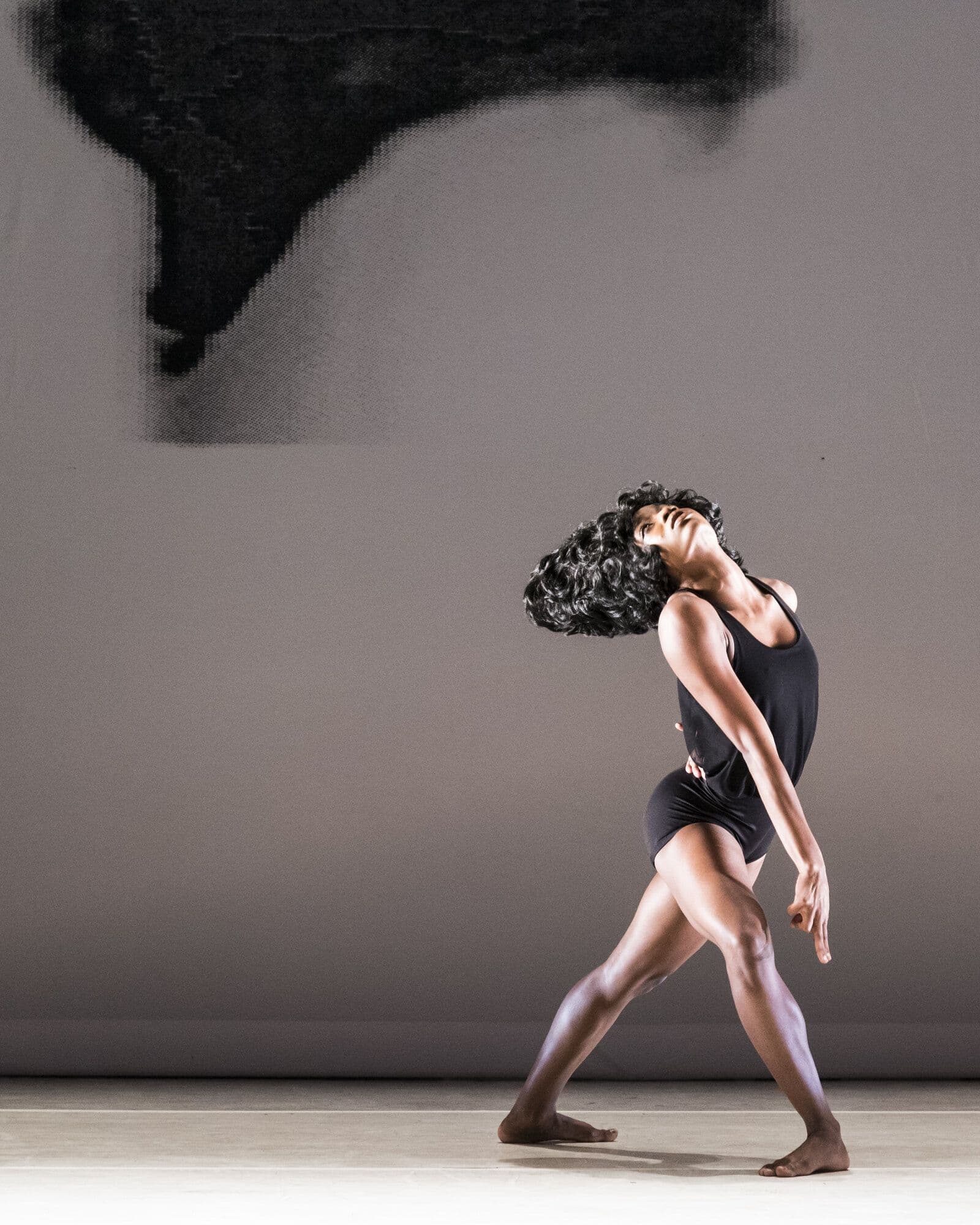 A dancer in a black top and shorts leans back with her head tilted skyward, dark curly hair swinging, one leg planted forward in a wide stance, set against a muted gray backdrop with a dark abstract shape overhead.