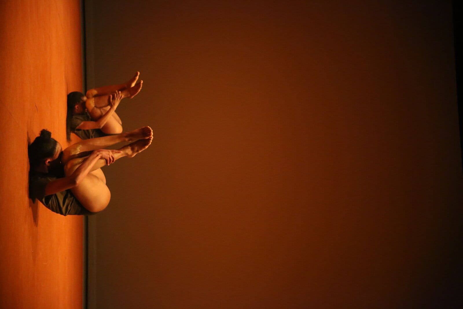 A dancer lies curled on the floor in a low, grounded pose with legs extended and gold-painted knees, gazing toward the camera under warm stage lighting.