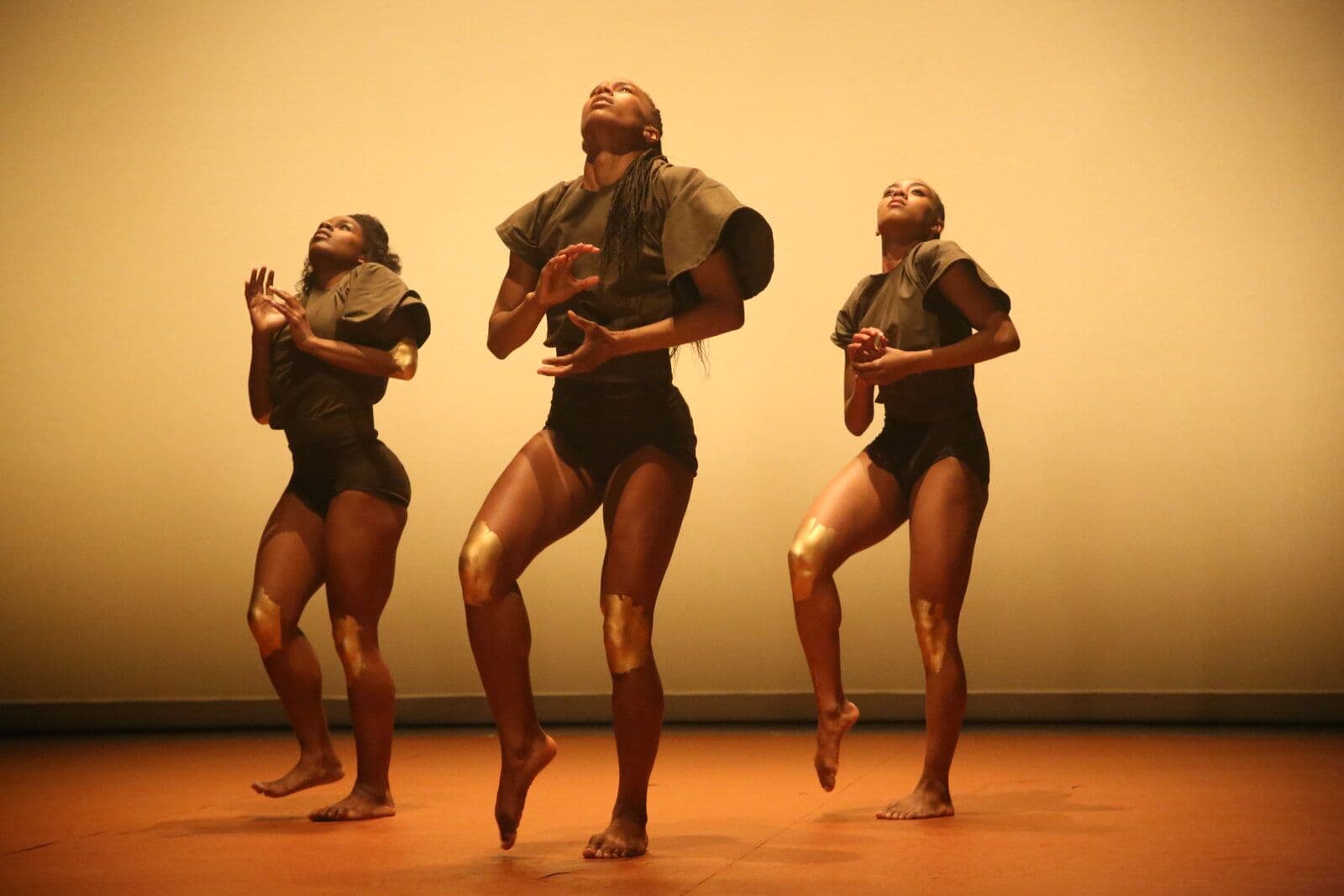 Three dancers perform in unison, reaching upward with bent knees and expressive hands, wearing olive tops and dark shorts with gold accents on their legs, lit warmly against a soft amber backdrop.