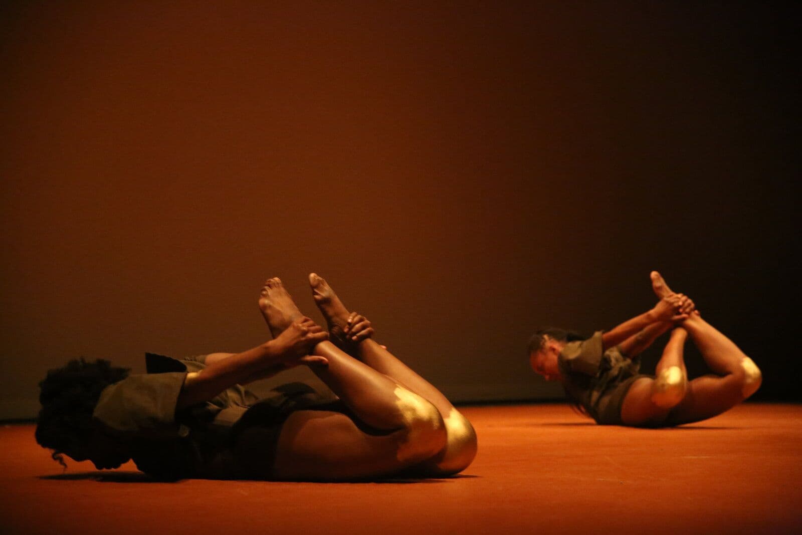 Two dancers lie curled on the floor with legs raised and hands clasped around their ankles, silhouetted in warm amber stage light.