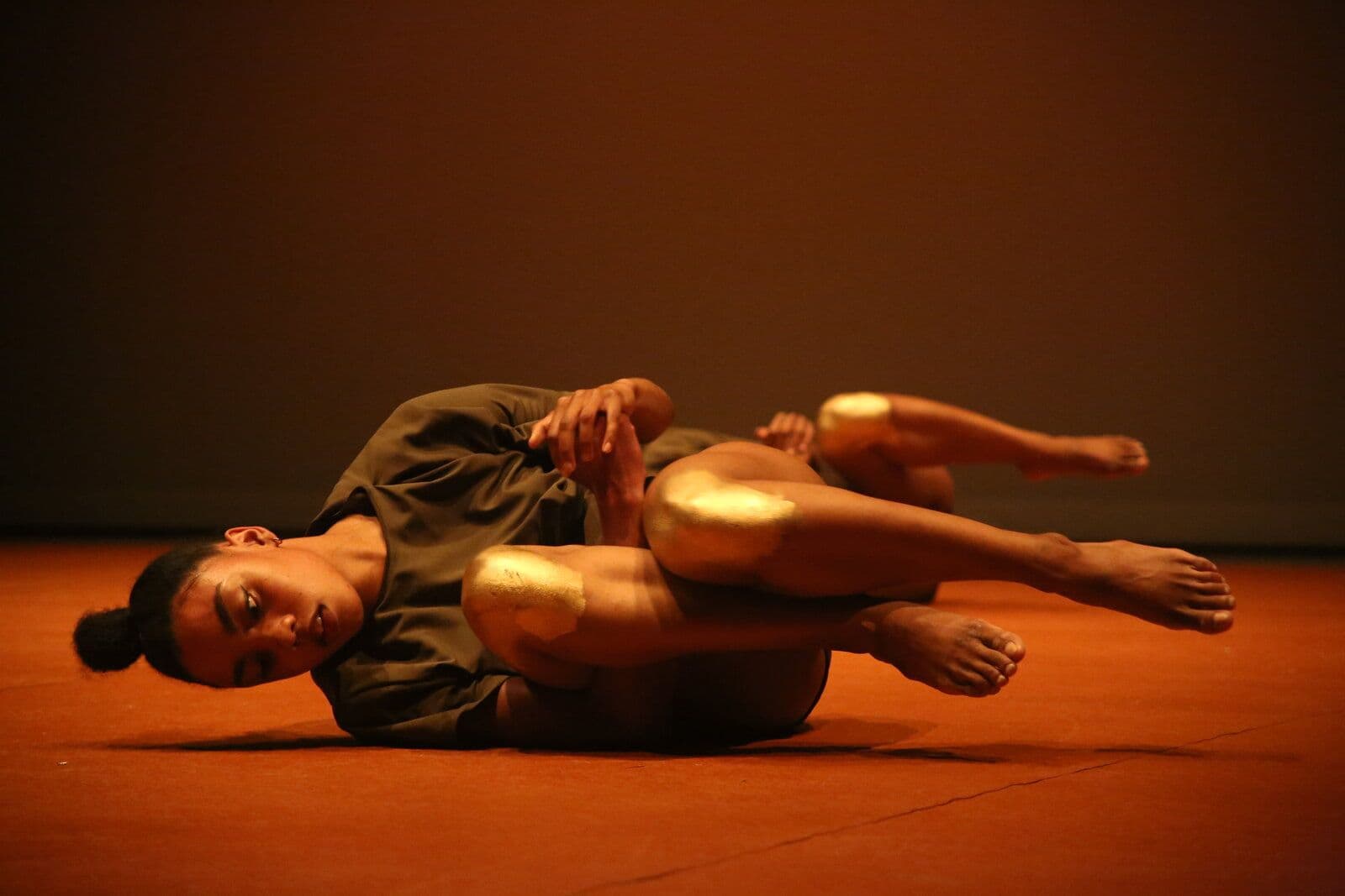 Two dancers perform matching curled floor poses, holding their raised feet, lit in warm amber tones against a dark stage.