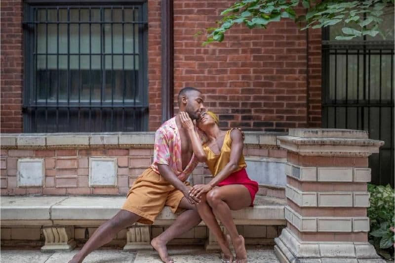Two dancers share an intimate, tender pose while seated on a stone bench outdoors in front of a brick building.