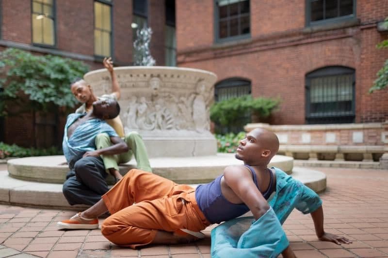 Three dancers perform expressive, fluid poses around an outdoor stone fountain in a courtyard.
