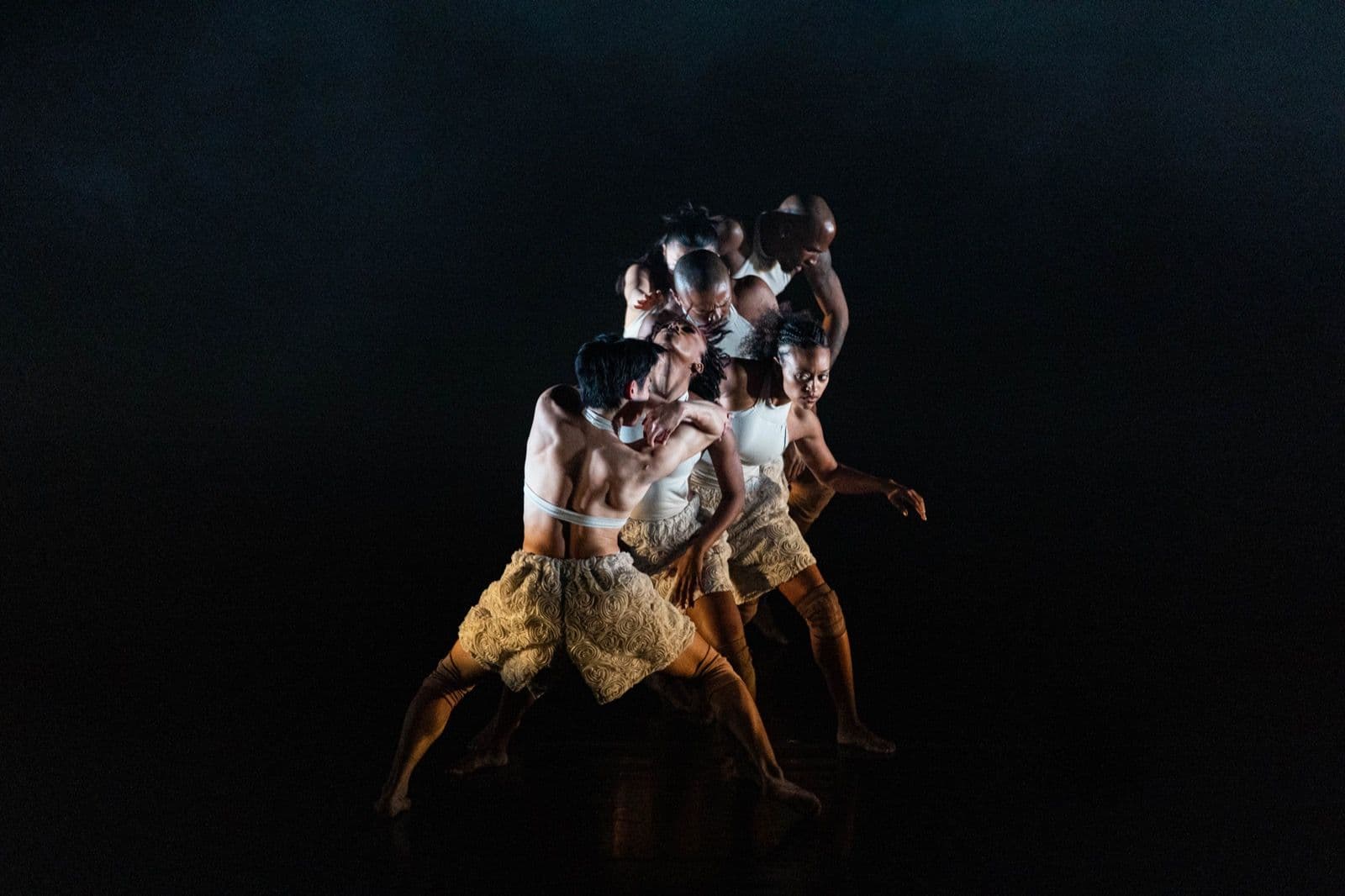 A group of contemporary dancers in white tops and textured shorts grouped tightly together under a dramatic dark spotlight.