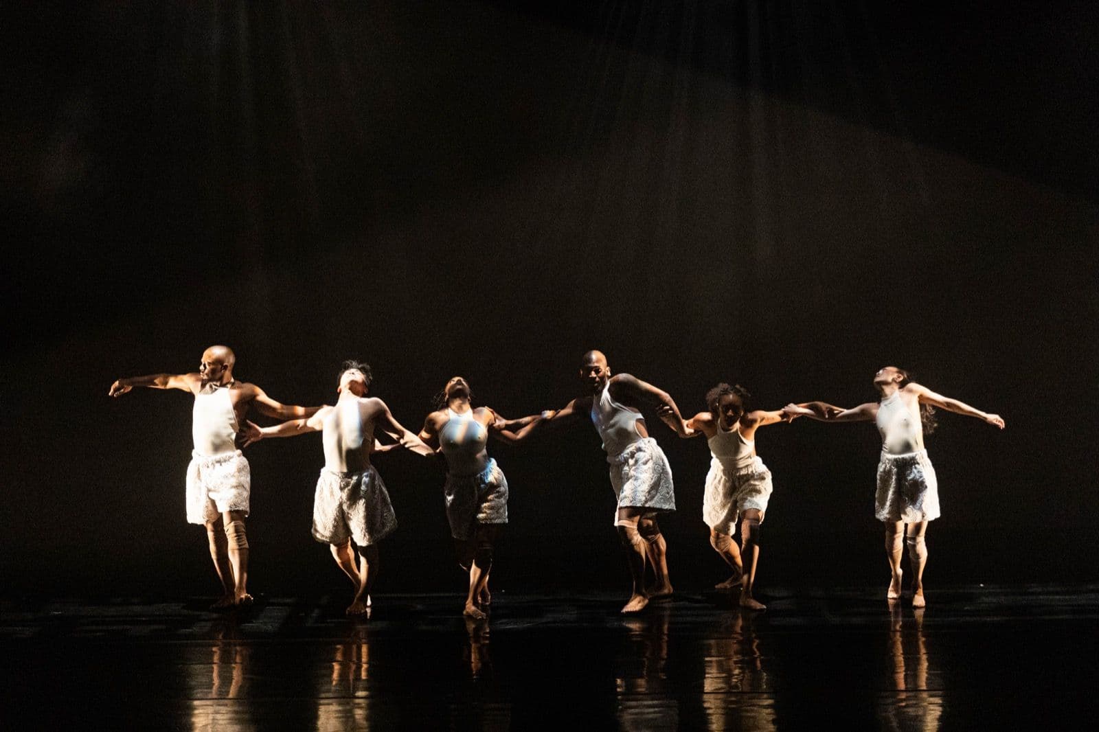 Six modern dancers in white outfits holding hands in a line across a dimly lit stage, leaning in expressive poses.