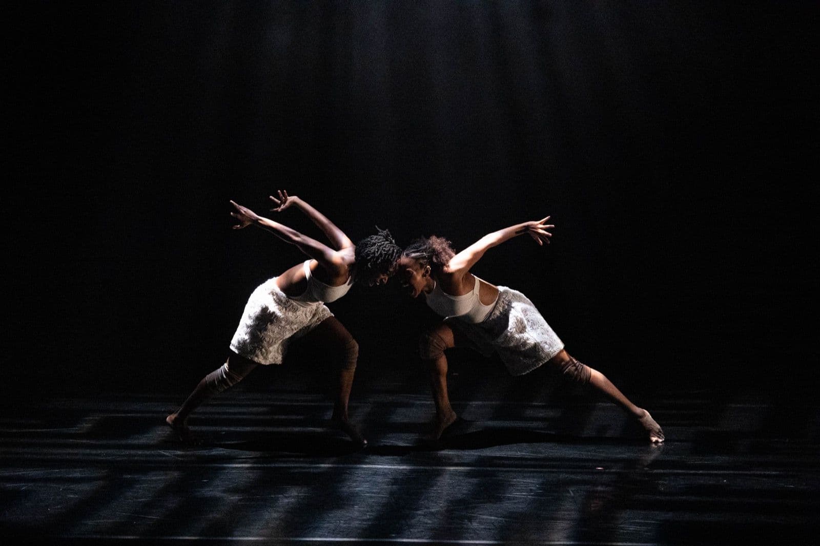 Two female contemporary dancers leaning towards each other with outstretched arms on a dark, moodily lit stage.