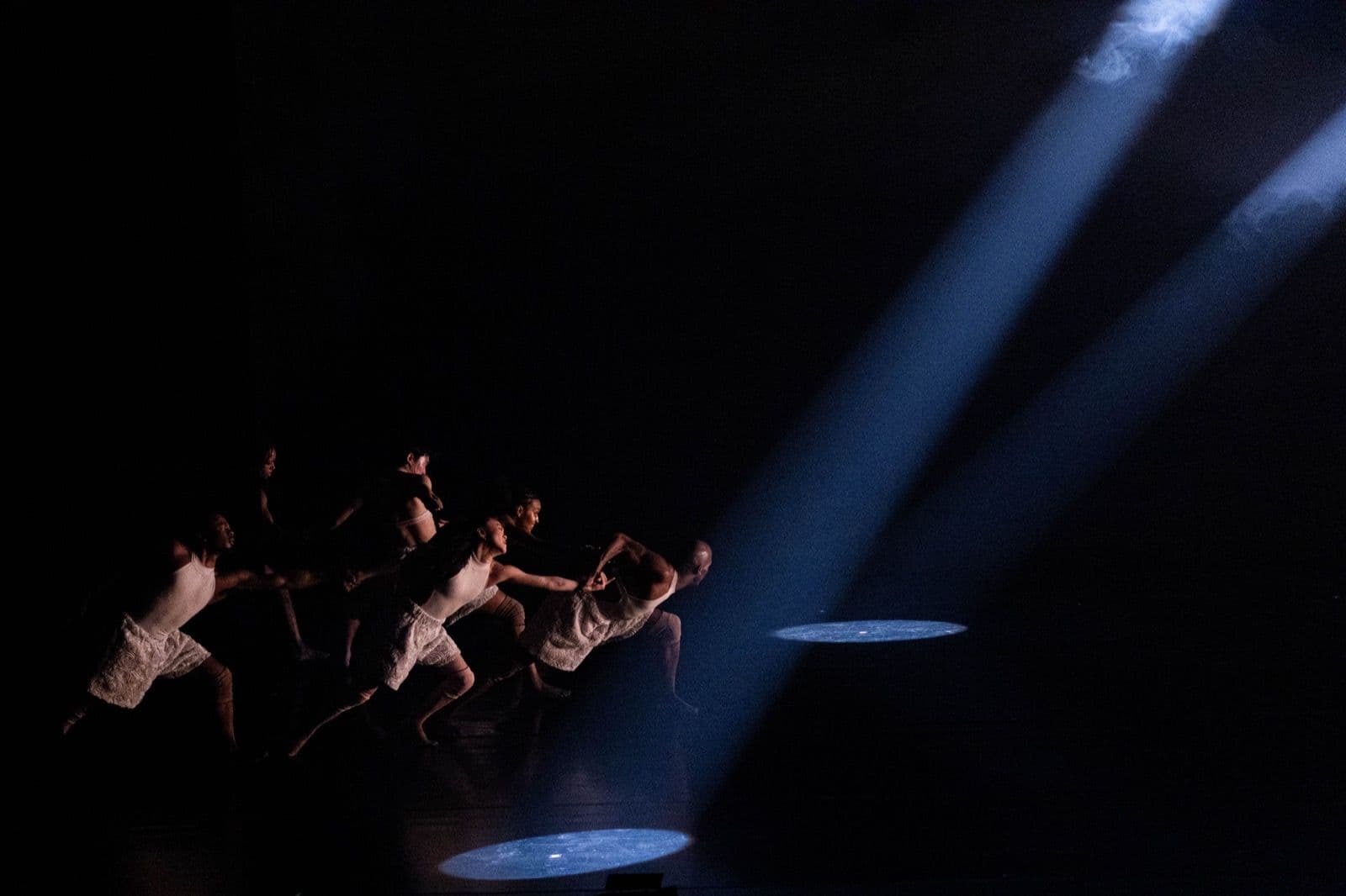 A group of dancers reaching towards two bright blue spotlights cutting through a dark stage.