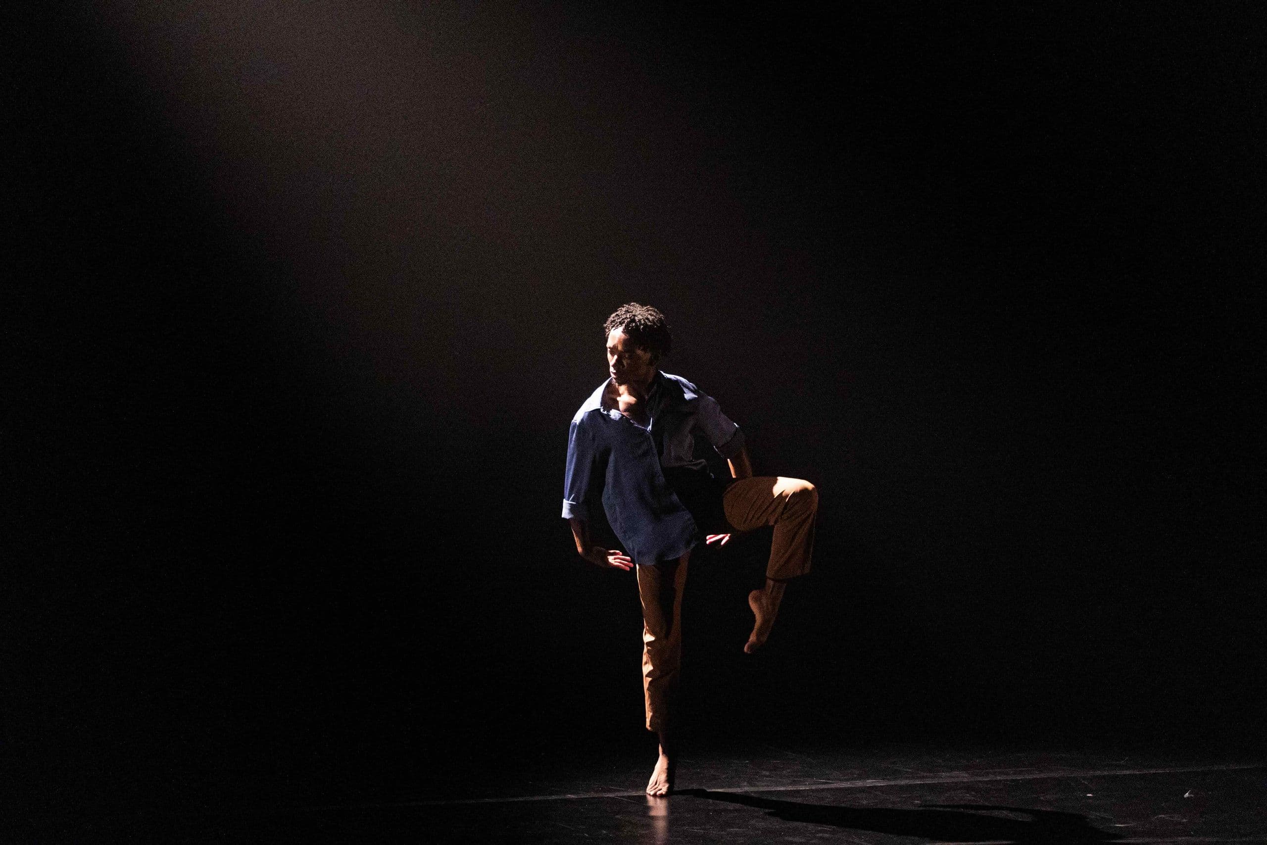 A solo male dancer wearing a blue shirt and tan pants performs barefoot on a dark stage illuminated by a single overhead beam of light.