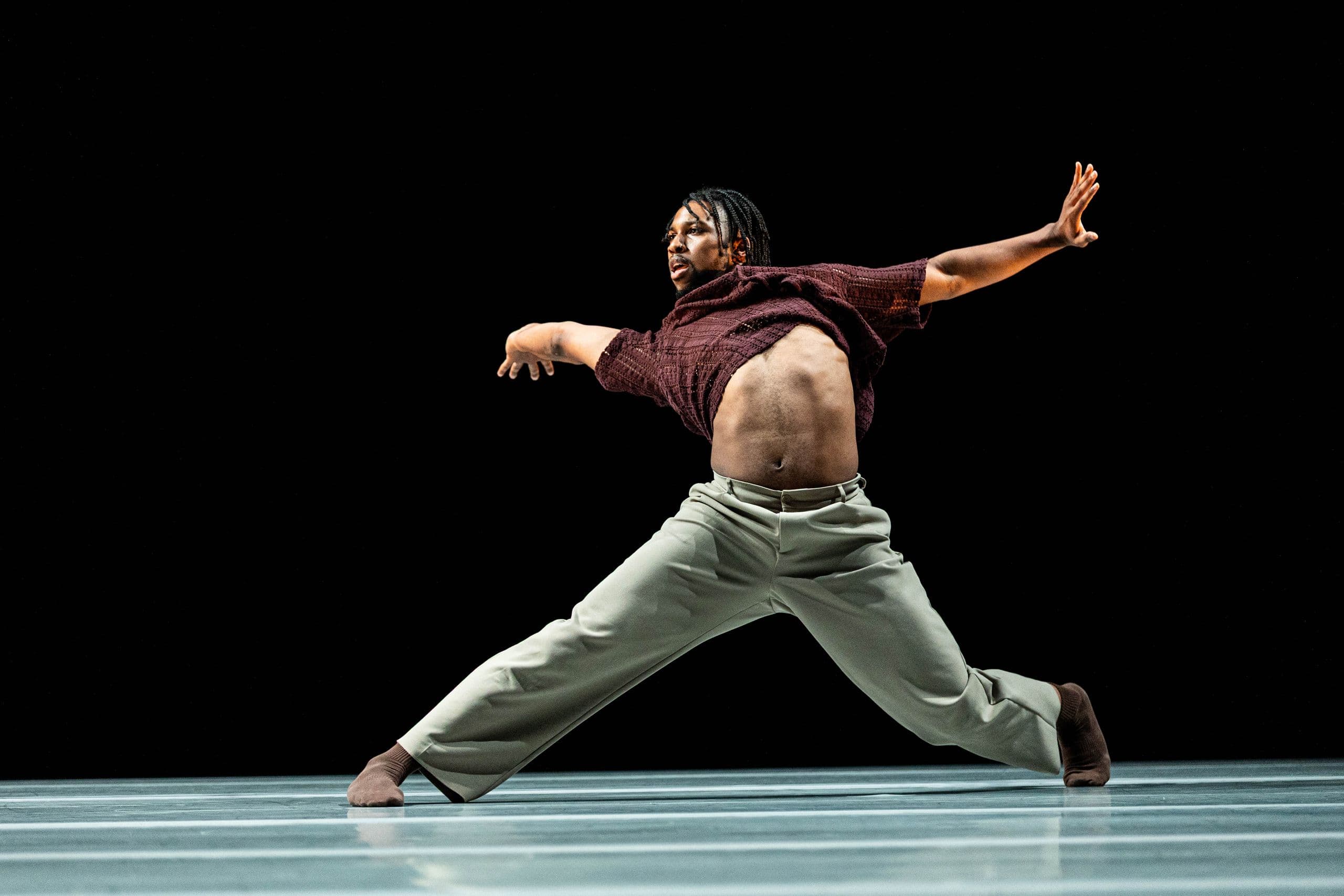 A male contemporary dancer in a wide stance on a dark stage, wearing brown socks, gray trousers, and a maroon crop top with arms stretched out wide.