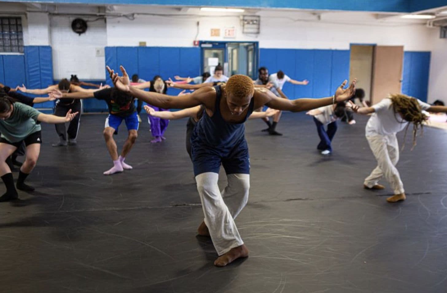 A group dance class in a studio with blue padded walls, dancers reaching their arms out in unison as an instructor in front leads the movement, leaning forward with arms spread wide.