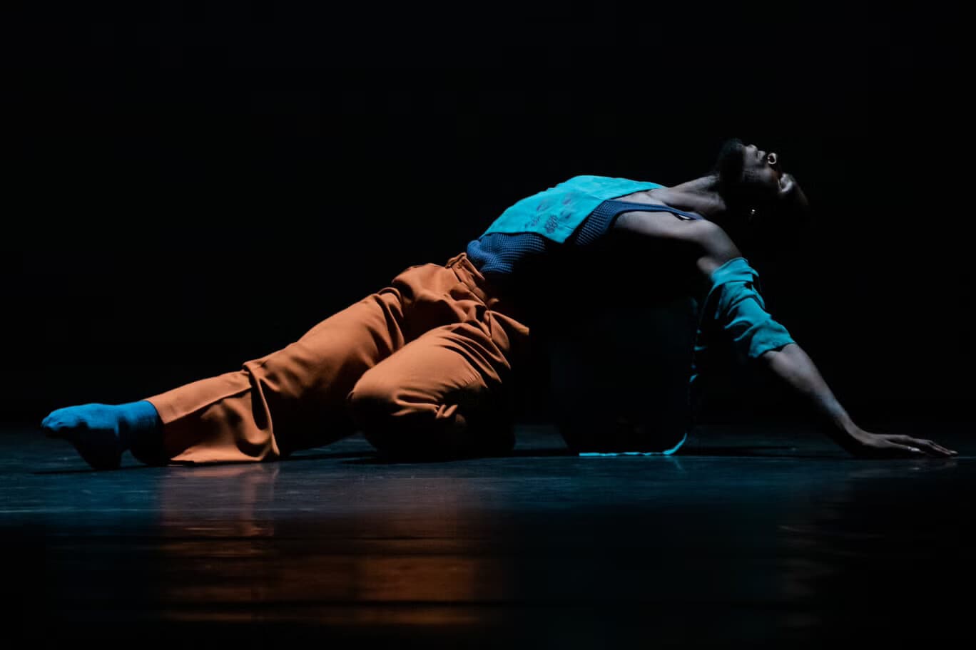 Dancer in a rust-orange and teal costume arched backward over a low platform, one arm reaching toward the floor, lit in deep blue against a black background.