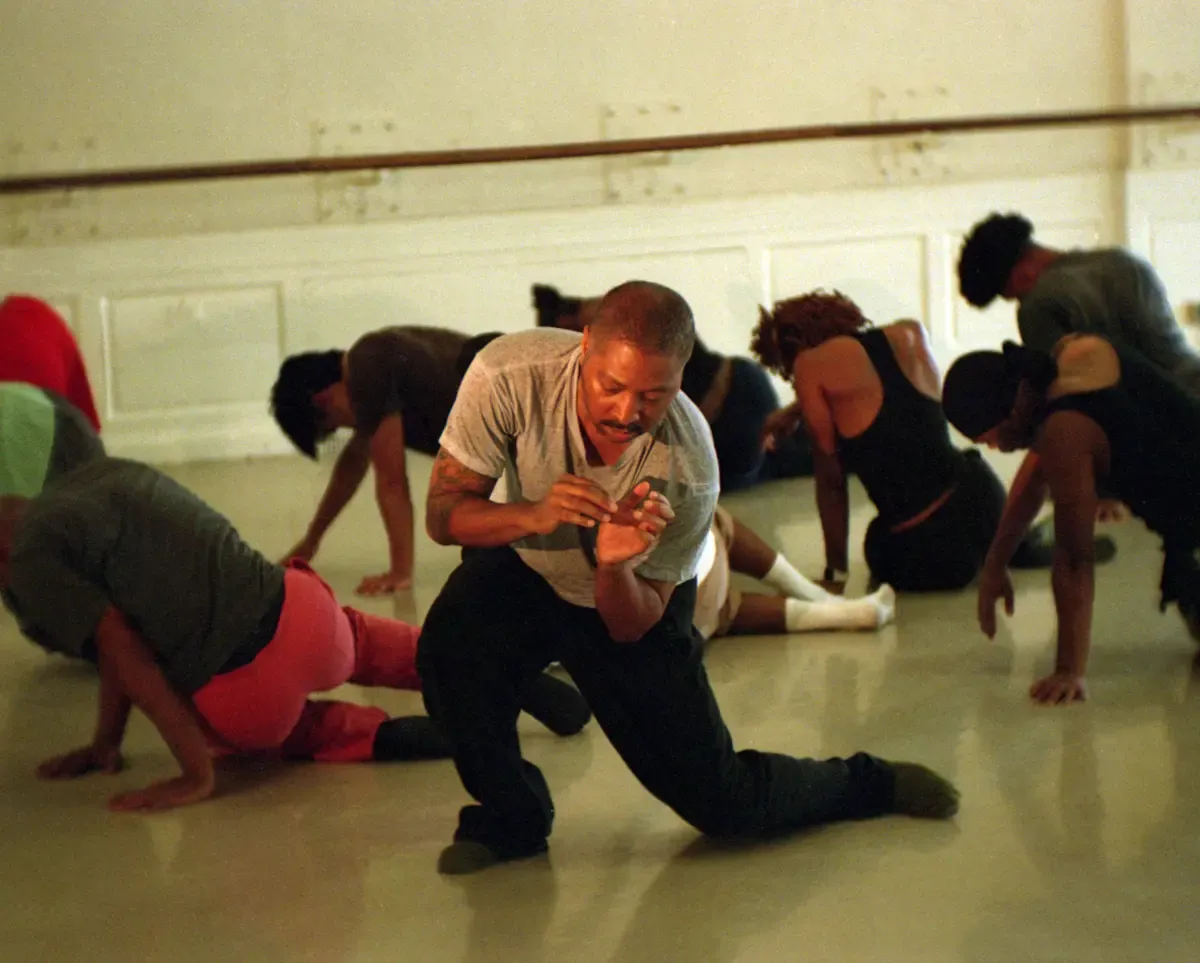 Kyle Abraham kneeling and demonstrating a movement in a studio, with several dancers behind him in various floor stretches near a ballet barre.